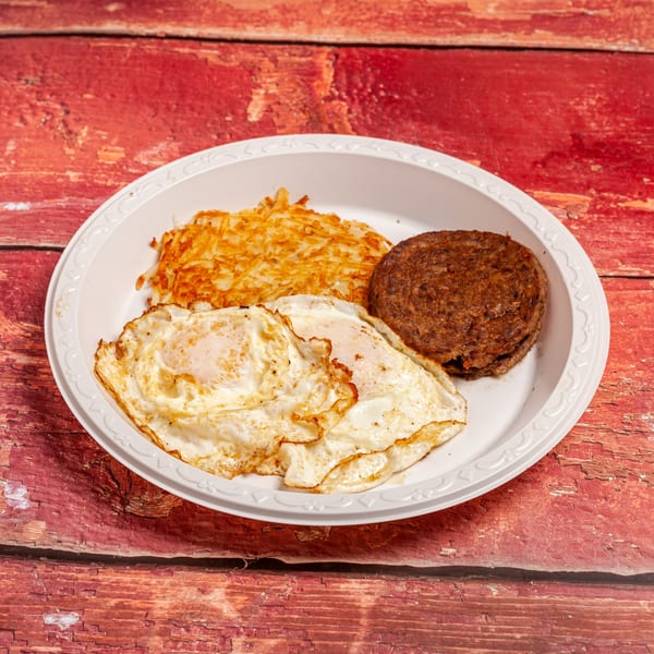 A white plate holds two fried eggs, hash browns, and a sausage patty on a red wooden surface.