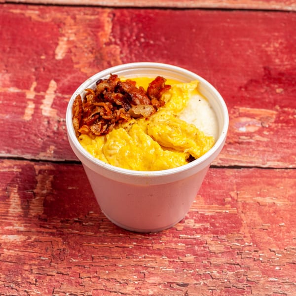 A white cup filled with scrambled eggs, grits, and crispy bacon bits on a red wooden surface.