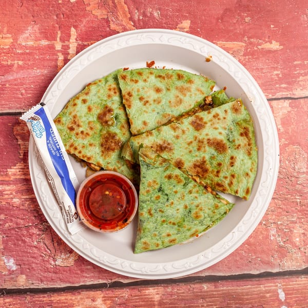 Green quesadilla slices with salsa and a sour cream packet on a white plate, on a red wooden surface.